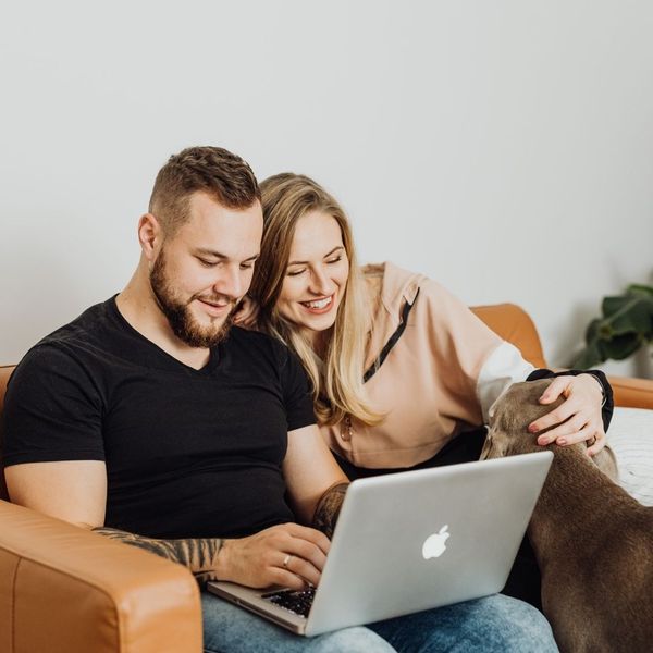A happy couple sitting on a couch, holding a laptop and smiling at each other. They appear to be planning a trip together. A happy couple sitting on a couch, holding a laptop and smiling at each other. They appear to be planning a trip together.