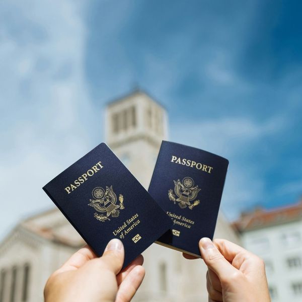 Two hands holding up blue American passports against a bright, cloudy sky with a historic building in the background. The image evokes international travel and adventure. Two hands holding up blue American passports against a bright, cloudy sky with a historic building in the background. The image evokes international travel and adventure.