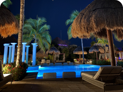 A luxurious resort pool surrounded by palm trees and tiki huts, beautifully illuminated at night.