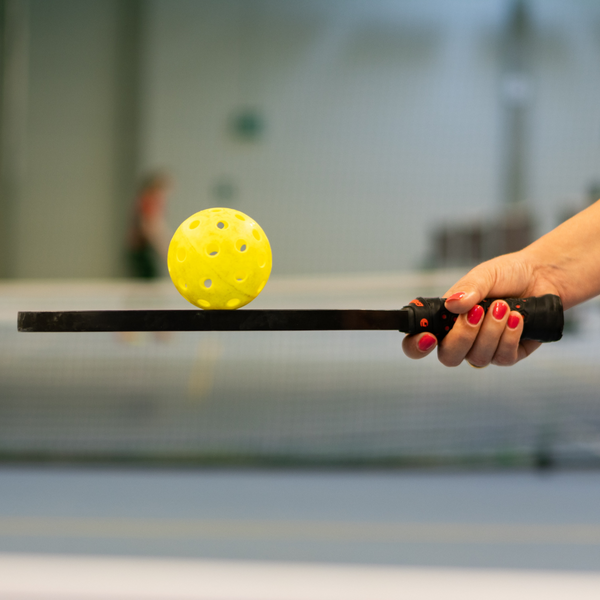 A person's hand holding a pickleball paddle with a bright yellow pickleball balanced on the surface. A person's hand holding a pickleball paddle with a bright yellow pickleball balanced on the surface.