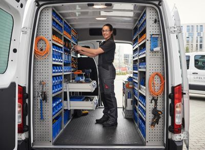 A smiling tradeswoman in safety glasses and work clothes organizes tools within the fully equipped interior of a white service van, featuring numerous storage bins and shelves. Tradeswoman Organizing Tools in Service Van