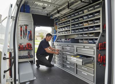 A technician kneels inside a white work van, accessing tools from well-organized drawers and shelves. The van features extensive storage solutions. Technician working in a fully equipped service van