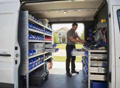 A technician works at a workbench inside a service van, fully outfitted with organized shelves, bins, and tools, with a house in the background. Service Van Interior with Technician and Organized Tool Storage