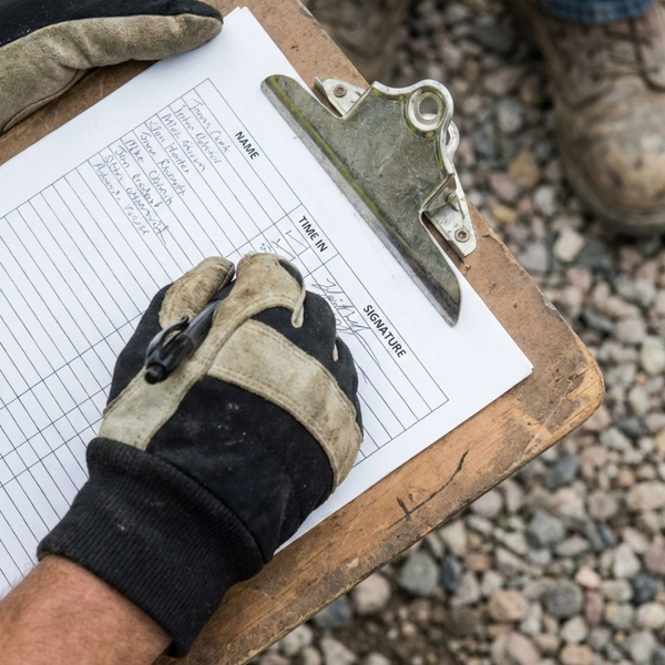 A close-up view of a safety officer's gloved hand checking names on a paper safety log clipboard at a work site.