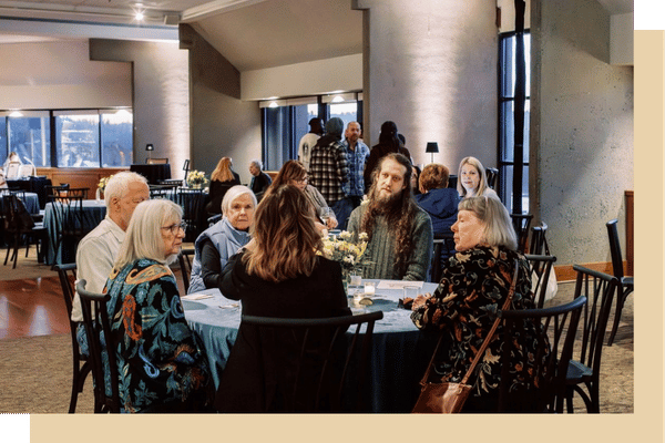 A banquet hall set with round tables covered in dark cloth, each with floral centerpieces and place settings, while guests sit and staff stand near a buffet or service area.