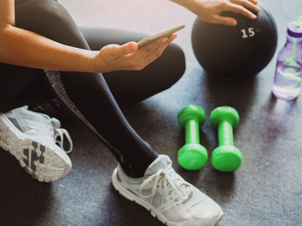 Image of a woman sitting on the floor with her smartphone, surrounded by workout equipment. 