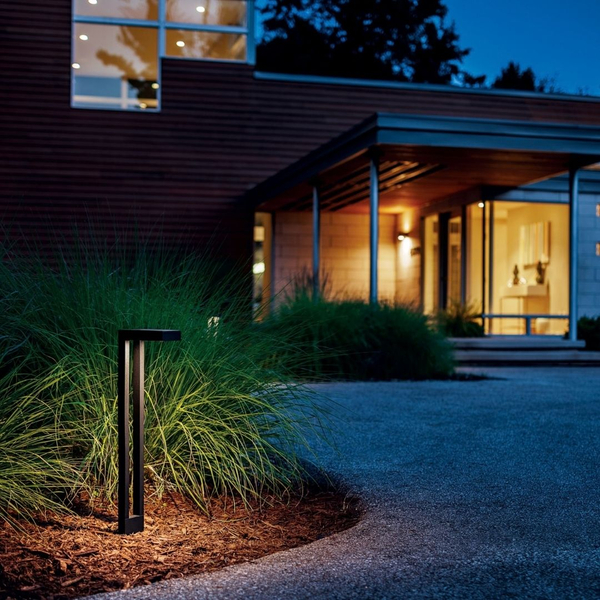 Modern black LED bollard landscape light illuminating a garden bed and driveway at dusk.   