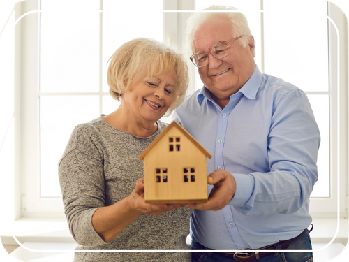smiling older couple holding out wooden house smiling older couple holding out wooden house