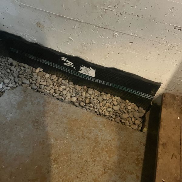 A close-up of a basement wall with a black drainage mat and gravel visible along the base.