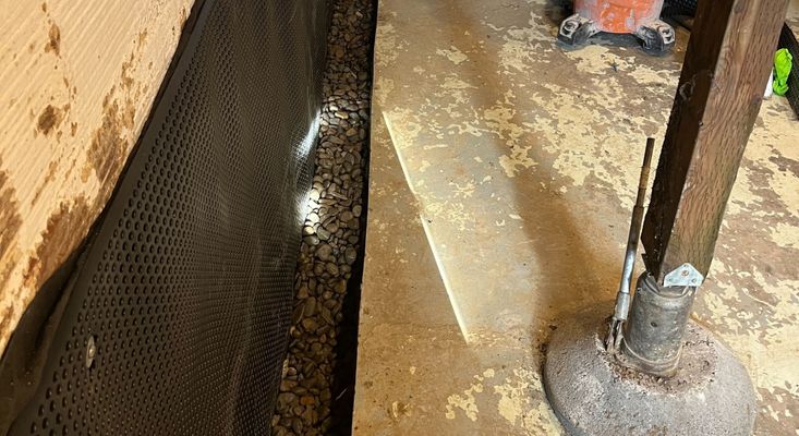 A section of a basement showing a black dimpled drainage membrane against the wall, with gravel at its base, and a support beam in the foreground.