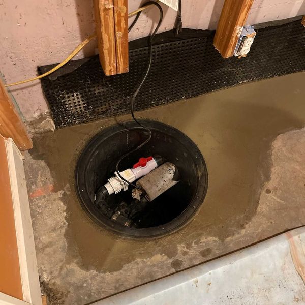 An overhead view of a sump pump installed in a basement, surrounded by concrete and a black dimpled membrane.