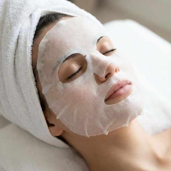 A woman relaxing with a hydrating sheet mask on her face and a towel on her hair in a spa settingA woman relaxing with a hydrating sheet mask on her face and a towel on her hair in a spa setting