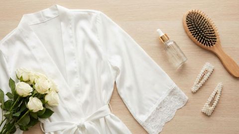 A flat lay of bridal beauty items including a white silk robe, white roses, a hairbrush, and a facial serum bottle A flat lay of bridal beauty items including a white silk robe, white roses, a hairbrush, and a facial serum bottle