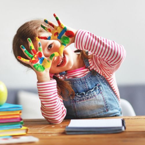 Child peeking through her hands, covered in colorful paint.