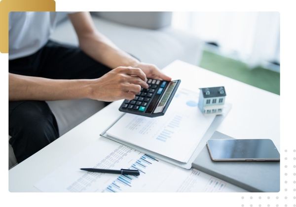 Close-up of a person using a calculator with financial documents, charts, a pen, and a small model house to represent financing options for home remodeling.