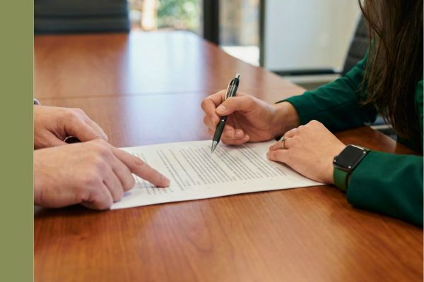 Close-up of an attorney in a green jacket and a client's hands reviewing a legal document together on a wooden table.