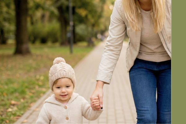 A blonde woman walking hand-in-hand with a young child on a paved path in a sunlit, blurred park setting.