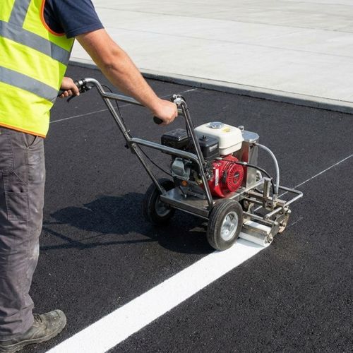 A high-angle view shows a worker precision-applying bright white thermoplastic striping onto dark cured asphalt with a hand machine.