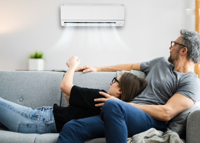 Two people relaxing on a couch while one turns on a wall-mounted AC unit with a remote control
