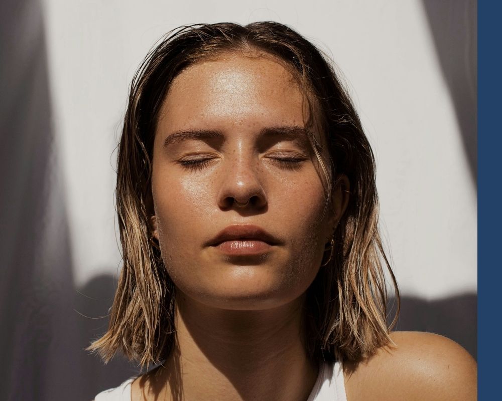 Close-up portrait of a woman with eyes closed and freckles, bathed in natural sunlight and shadows, representing a serene and natural aesthetic.