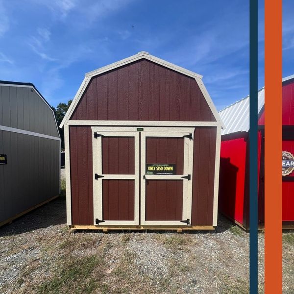 A brown shed with off-white trim and black doors, featuring a gambrel roof, set on a gravel lot under a partly cloudy sky.