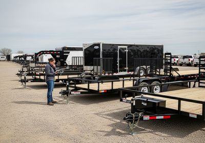 A woman inspects a trailer lot, reviewing documents while standing among various trailers, including flatbed and enclosed models, with pickup trucks visible in the background. Trailer Lot Inspection