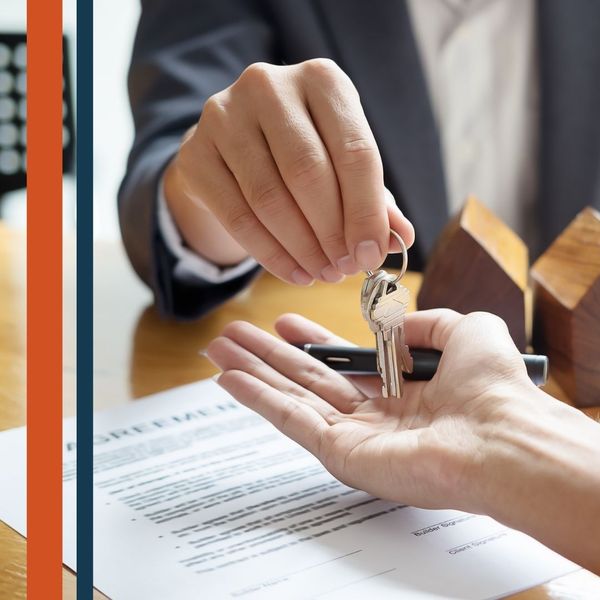 A person's hand extending a set of house keys towards another person's open hand, with a partial view of a contract and small house models on a desk.