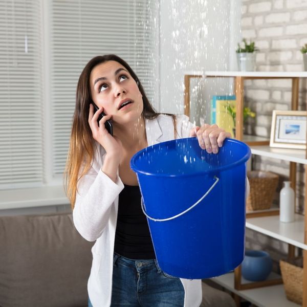 woman calls plumber while using bucket to catch leaking pipes