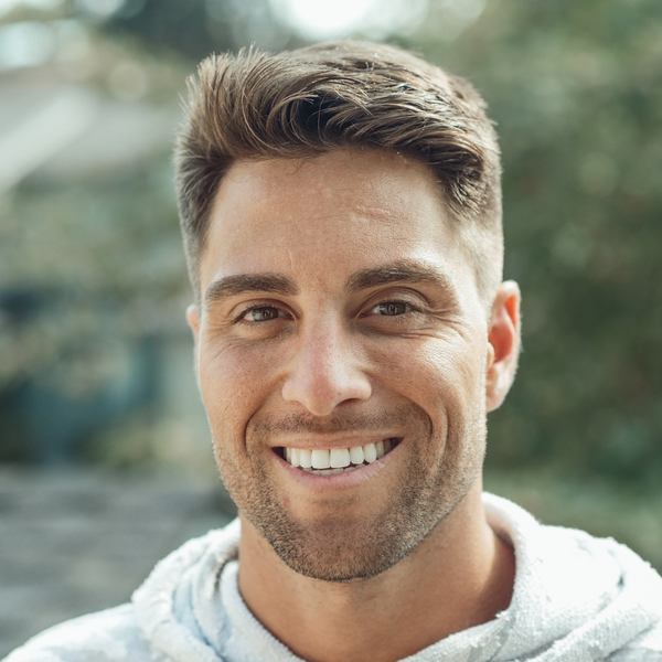 A close-up photograph of a man with a short beard smiling, wearing a white textured hoodie, outdoors.