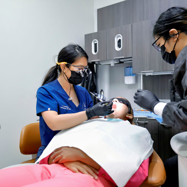 A dentist and an assistant in protective gear are treating a patient reclining in a dental chair in a clinical room.