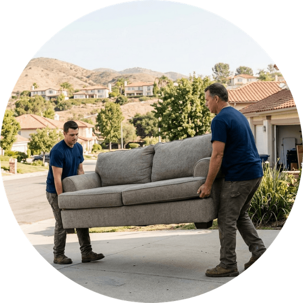 Two unbranded crew members in matching blue t-shirts carefully carrying a large fabric sofa down a residential driveway in Westlake Village, California, with foothills and Spanish-style homes visible in the background.