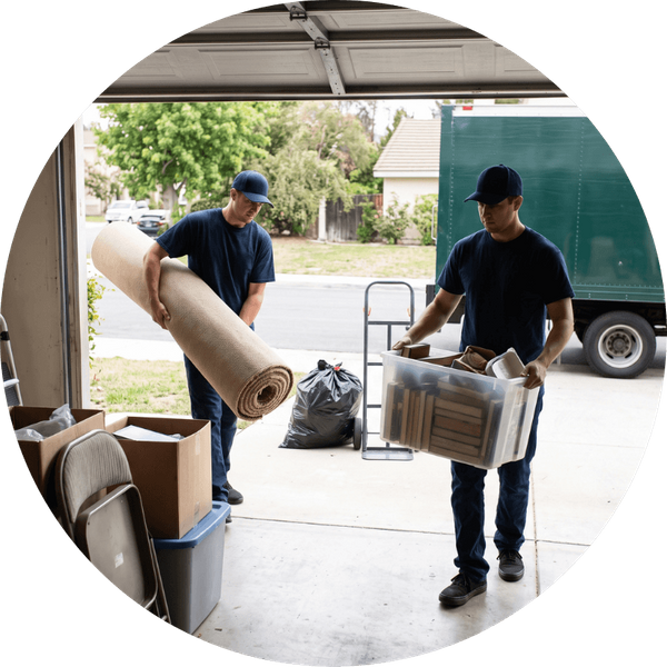 Two junk removal workers in blank dark blue shirts and caps carry bulky items—one lifting a rolled-up tan carpet and the other carrying a plastic storage tub filled with discarded materials—out of an open garage toward an unbranded green truck parked in a 