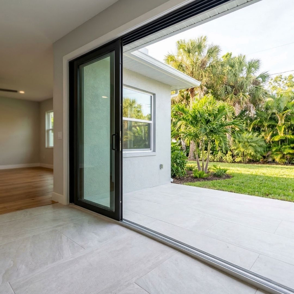 An inviting interior space connects a room with a natural wood floor to a bright outdoor patio through large, black-framed sliding glass doors.