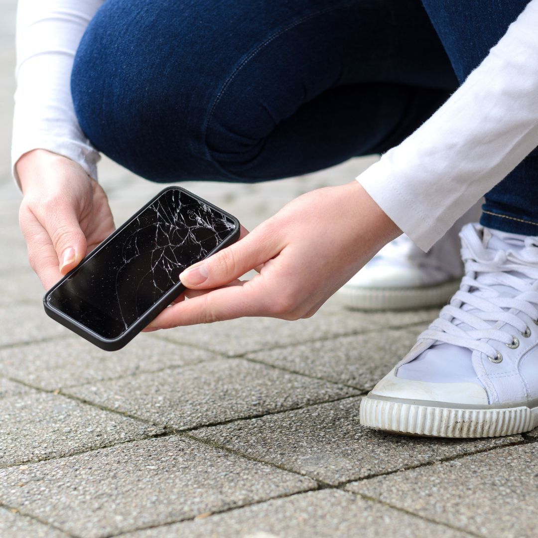 Person kneeling down to pick up a cell phone with a cracked screen. 