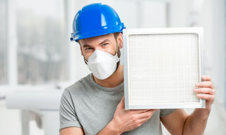 Person wearing a hard hat and mask, and holding up a home air filter