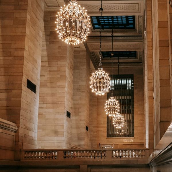 An upward shot inside a grand historical building, showing three ornate, illuminated chandeliers hanging from a high, decorative ceiling.
