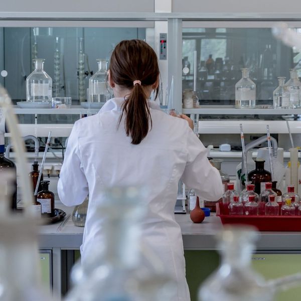 A close-up, over-the-shoulder view of a female scientist in a white lab coat working at a laboratory bench with various beakers and scientific equipment.
