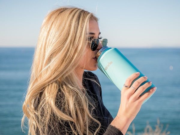 image of a blonde woman with sunglasses drinking from a stainless steel water bottle on the beach
