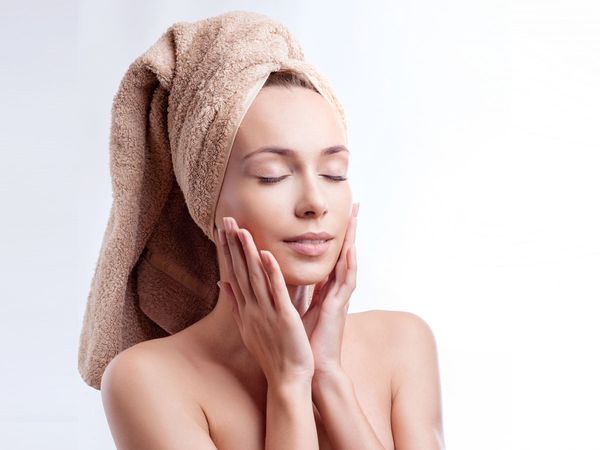 Woman wearing a towel around her head touching her face with both of her hands to show off her flawless skin