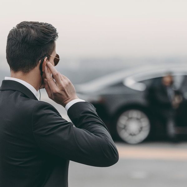 a man in a suite talking on an in ear device while someone gets out of a car in the background