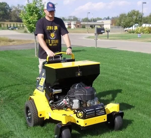Lawn care technician wearing a baseball cap and company T-shirt pushing a professional yellow and black fertilizer or seed spreader across a lush lawn.