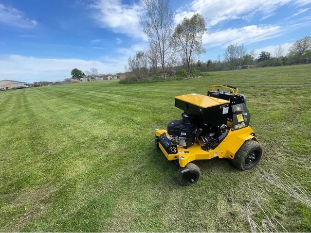 Yellow commercial lawn fertilizer spreader or lawn care equipment treating a large, freshly striped green lawn on a sunny day.