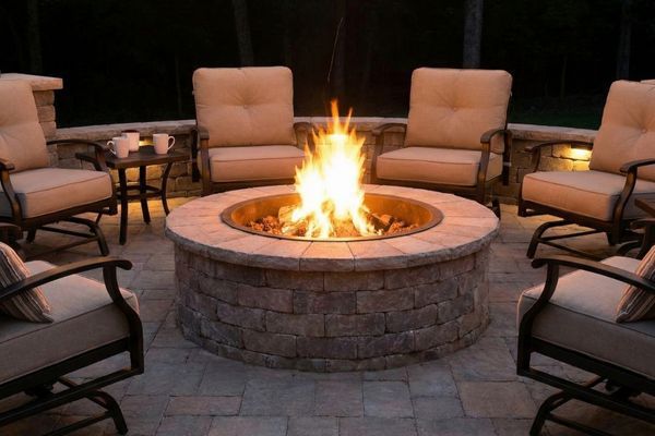 Lit custom stone fire pit on a paver patio at night, surrounded by comfortable seating and string lights.