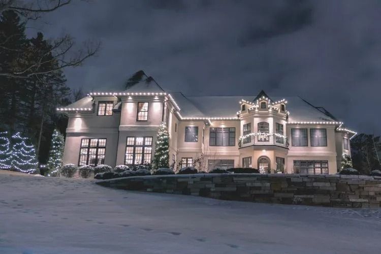Luxury home exterior decorated with bright white string lights along the rooflines and trees, covered in snow on a dark winter night.