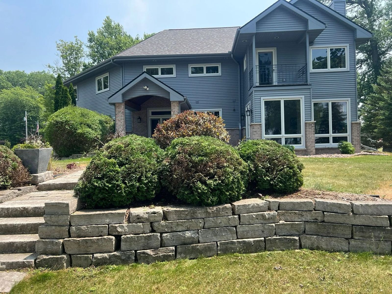 Large, modern blue-sided house with a stacked stone retaining wall and steps leading up to the front entrance, featuring large, mature green shrubs.