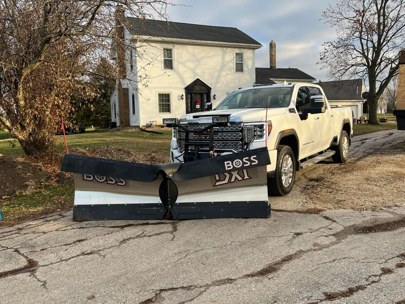 Commercial white pickup truck with a two-tone silver and black Boss snow plow blade attached, parked on a residential driveway in front of a white farmhouse.