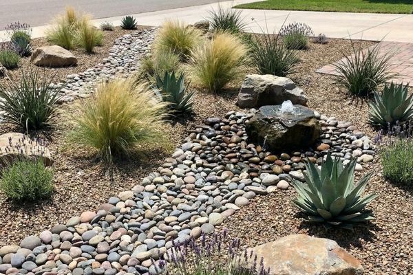 Low-maintenance xeriscape garden with river rocks, drought-tolerant plants like lavender, and a small bubbling rock water feature.