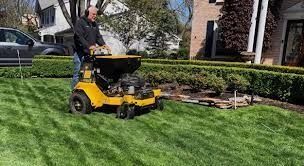 Professional lawn care technician operating a yellow commercial spreader/sprayer machine on a thick, striped green lawn next to a landscaped hedge.
