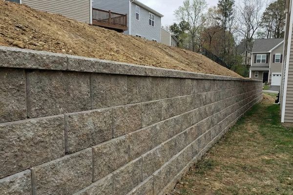 Tall, engineered block retaining wall holding back a hillside, with an integrated drainage pipe visible at the base.