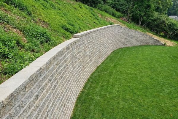 Tall, curving engineered block retaining wall holding back a steep hill to create a flat green lawn.